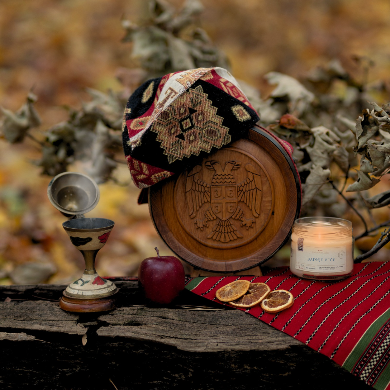 Mirisna sojina sveća Badnje Veče iz kolekcije Ususkani Dani brenda Dunavski Plamen – ručno pravljena sveća inspirisana Badnjakom, božićnim običajima i toplinom doma, prikazana uz jabuku, drvenu ikonu i prazničnu dekoraciju.