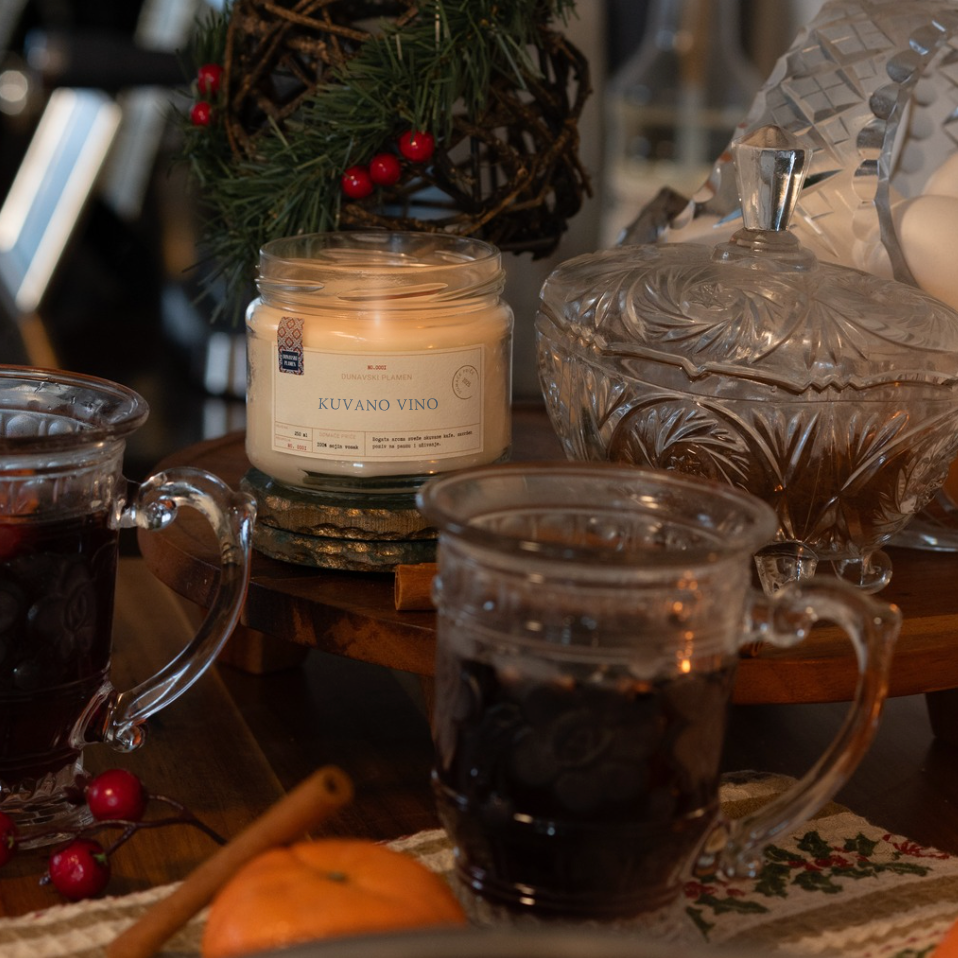 Mirisna sojina sveća Kuvano Vino iz kolekcije Ususkani Dani brenda Dunavski Plamen – ručno pravljena sveća sa toplim mirisima kuvanog vina, cimeta i pomorandže, okružena prazničnim ukrasima i kristalnim čašama.