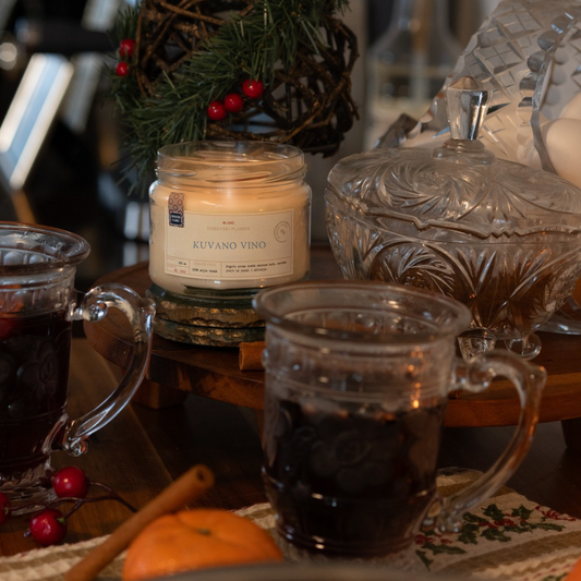 Mirisna sojina sveća Kuvano Vino iz kolekcije Ususkani Dani brenda Dunavski Plamen – ručno pravljena sveća sa toplim mirisima kuvanog vina, cimeta i pomorandže, okružena prazničnim ukrasima i kristalnim čašama.